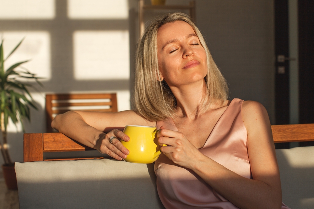 Relaxed Woman Enjoying Sunshine with a Yellow Mug - Annual Wellness Visit A blonde woman in a pink satin top smiles while holding a yellow mug, basking in warm sunlight indoors - Annual Wellness Visit