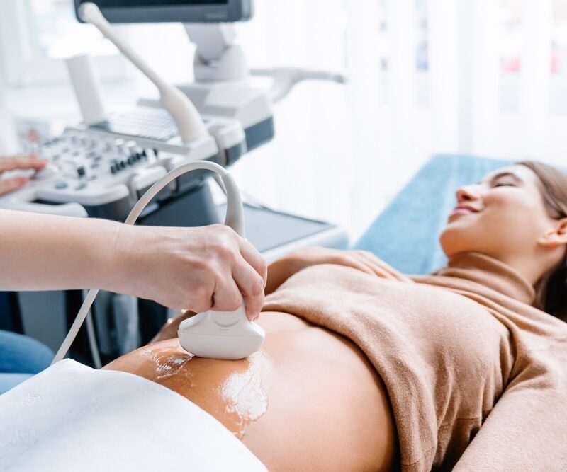 A smiling pregnant woman lies on an examination bed while a healthcare provider conducts an abdominal ultrasound - Ultrasound