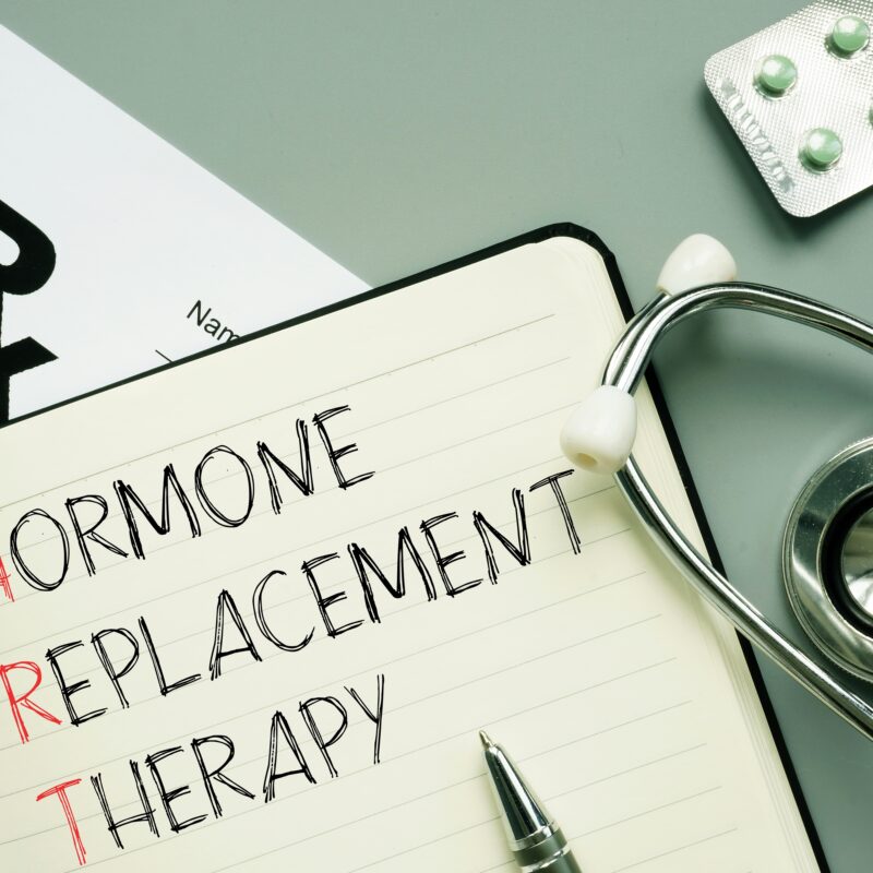 A notebook displaying the words "Hormone Replacement Therapy" alongside a stethoscope, pills, and a prescription note - HRT