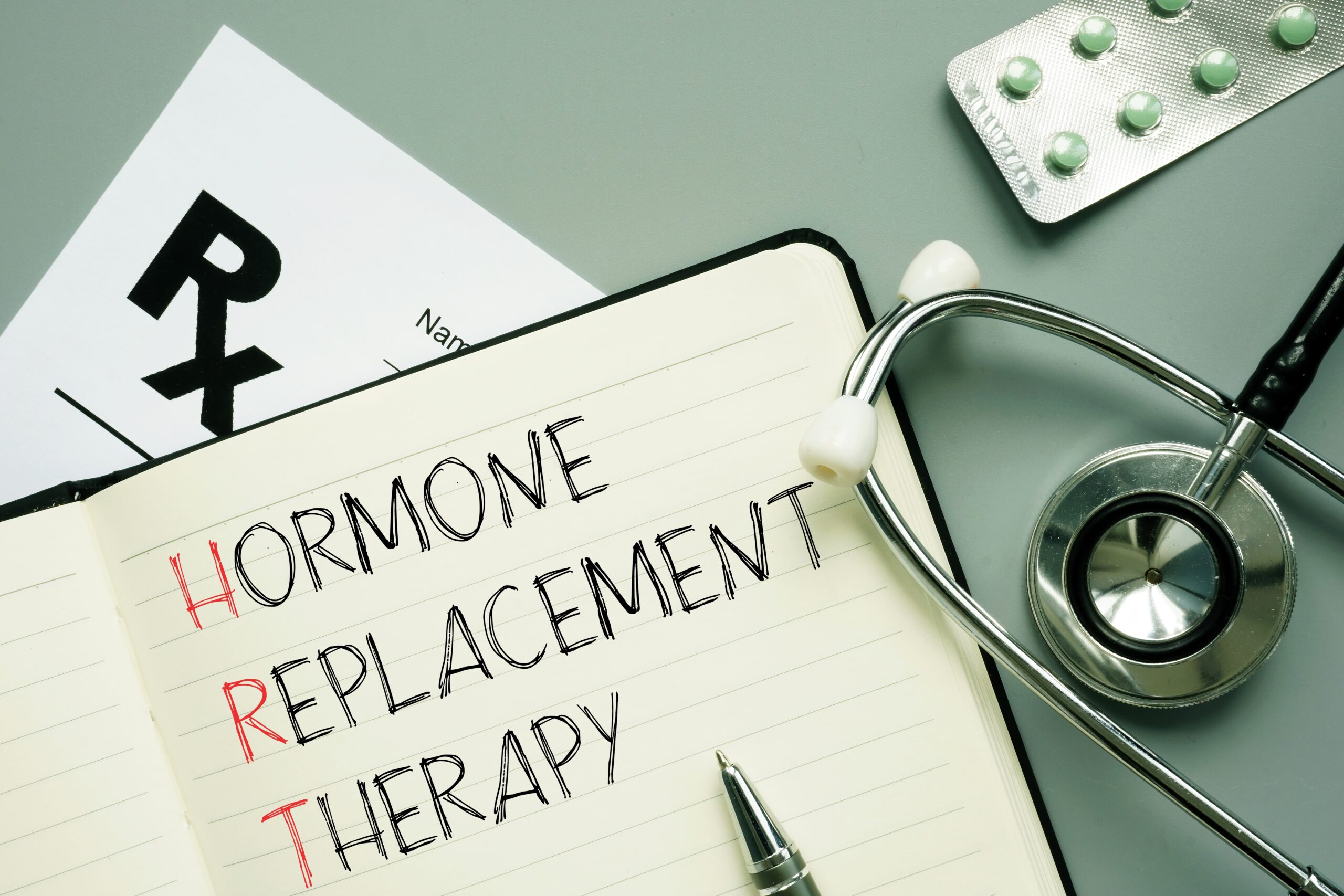 A notebook displaying the words "Hormone Replacement Therapy" alongside a stethoscope, pills, and a prescription note - HRT