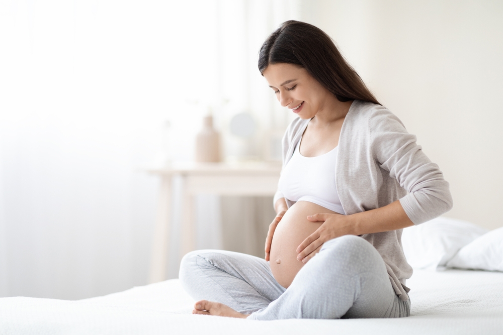 Smiling young pregnant woman sitting on bed in white bedroom - OBGYN in Milwaukee, WI Smiling young pregnant woman sitting on bed in white bedroom - OBGYN in Milwaukee, WI