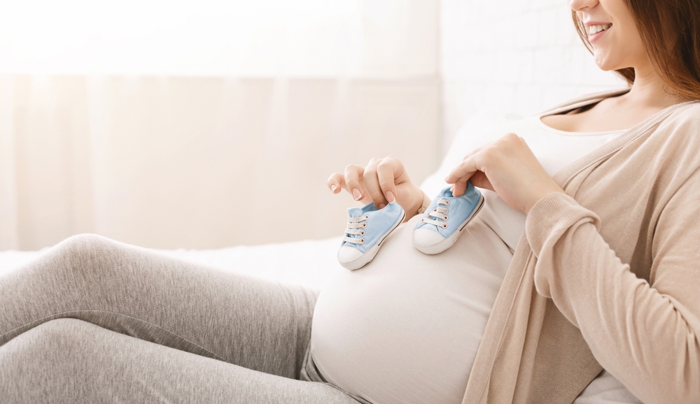 A pregnant woman holding small blue baby shoes on her belly, smiling in anticipation - Physicians for Women - Melius Schurr A pregnant woman holding small blue baby shoes on her belly, smiling in anticipation - Physicians for Women - Melius Schurr
